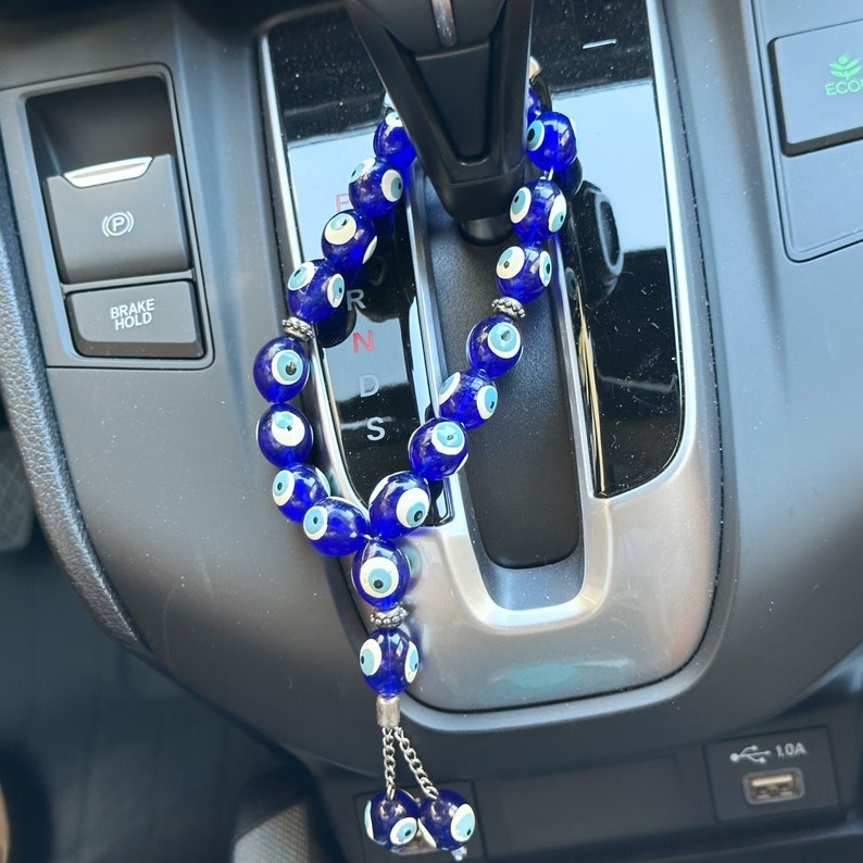 Op de afbeelding: Een blauwe kralen auto-hanger met meerdere glazen kralen, elk met een wit en blauw oogontwerp. De hanger hangt in een auto, met een zilveren ketting en een kwastje van kleinere oogkralen.