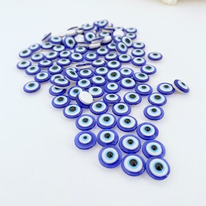 May include: A large group of blue evil eye charms with white and black centers. The charms are scattered on a white surface.