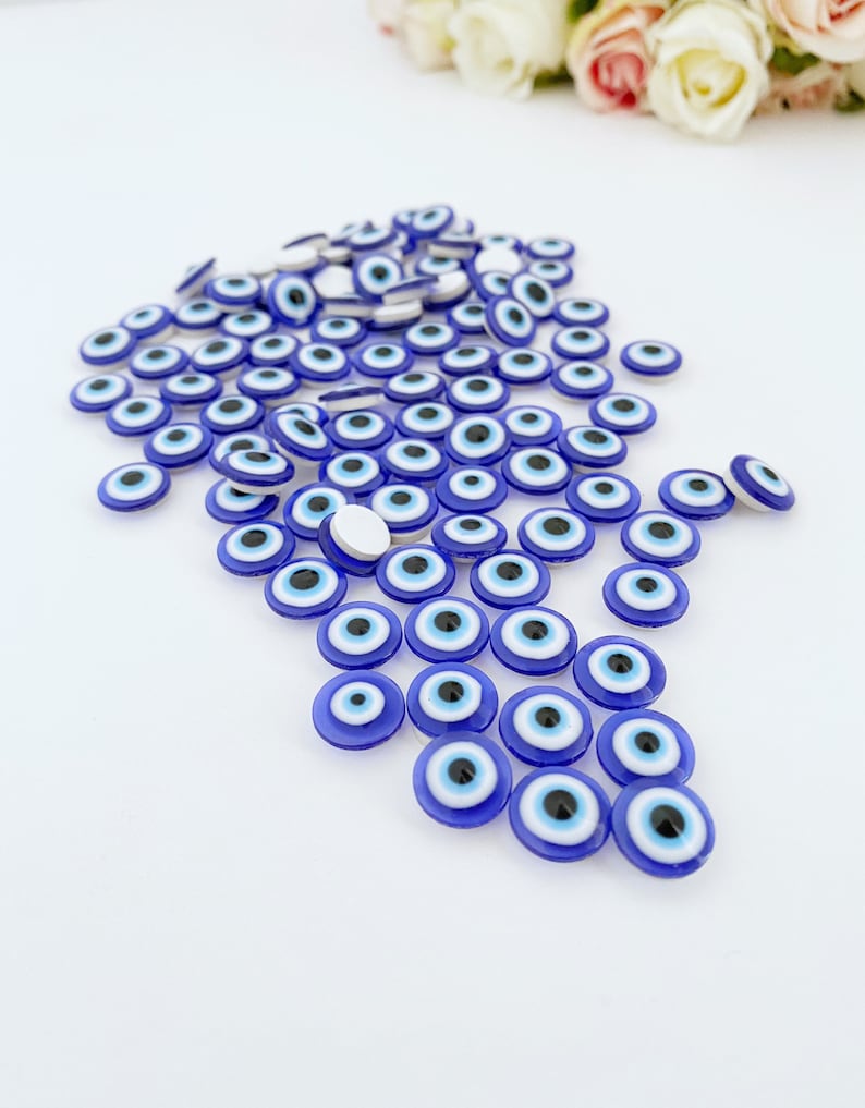May include: A large group of blue evil eye charms with white and black centers. The charms are scattered on a white surface.