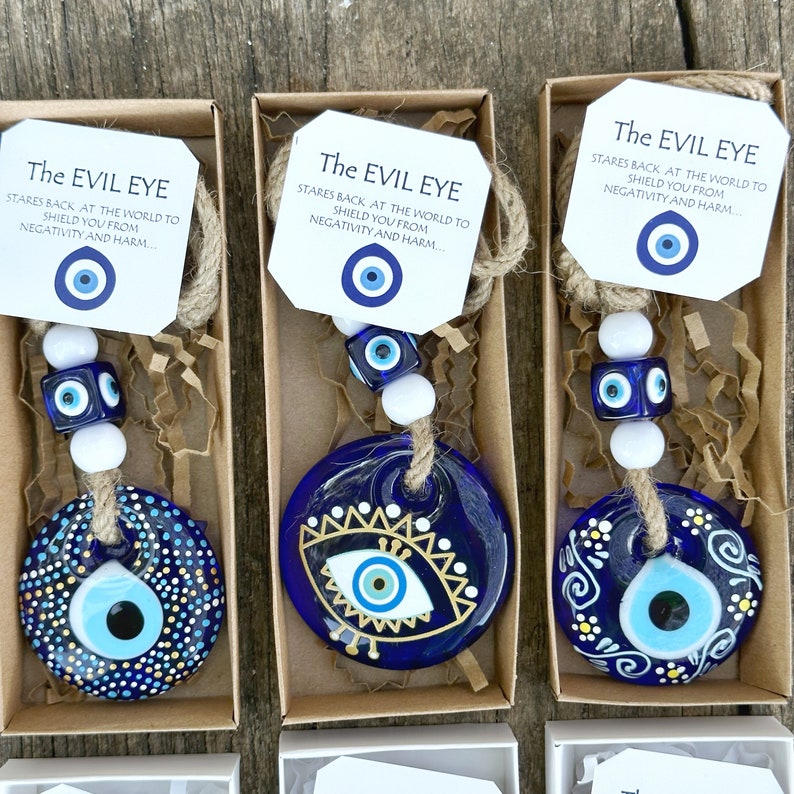 May include: Three small boxes each contain a blue glass evil eye charm with a white string and beads. The charms are decorated with gold paint and white dots. The boxes have a brown paper exterior and a white card with the text "The Evil Eye" and a description of the charm.