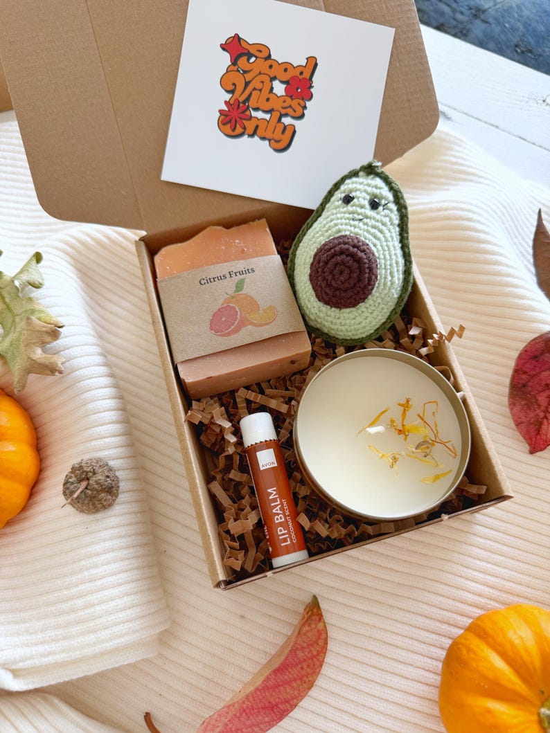 May include: A cardboard gift box containing a bar of Citrus Fruits soap, a crocheted avocado plush, a round tin candle with dried petals, and an Avon coconut scent lip balm. A card inside the lid features the text Good Vibes Only.