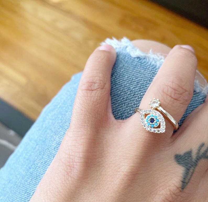 May include: Gold ring with a blue and turquoise evil eye charm and a smaller clear stone charm. The ring is on a finger wearing a blue denim jean.