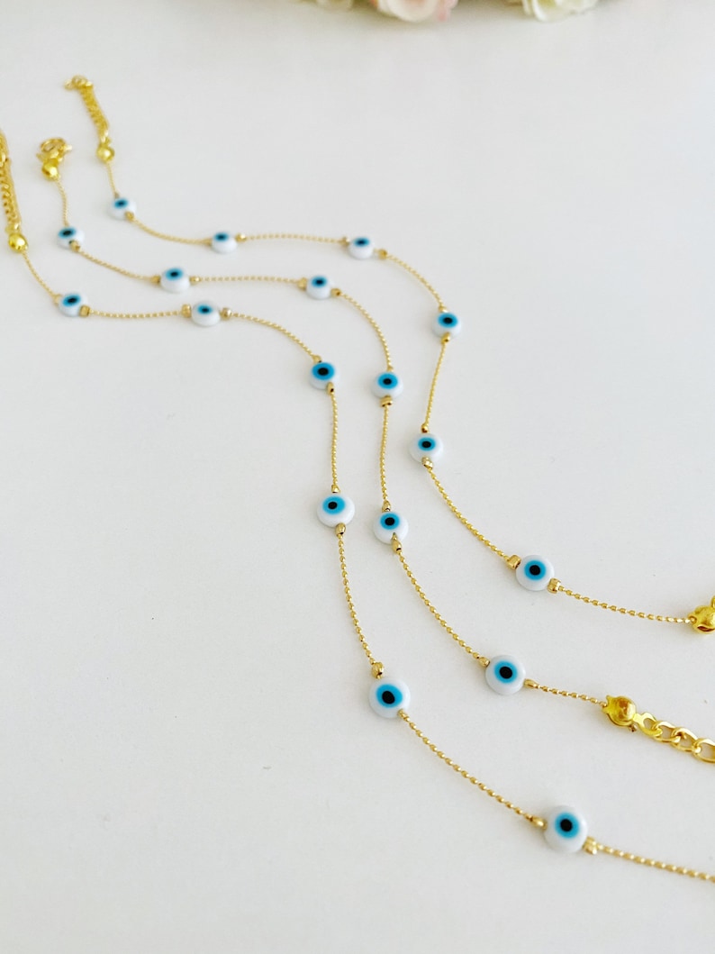 May include: Three gold chains with blue and white evil eye charms. The chains are layered on a white surface.