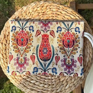 May include: A rectangular clutch with a vibrant floral pattern in red, orange, blue, and purple on an off-white background. The clutch has a zipper closure and a white cord handle. The bag is placed on a woven, circular surface.