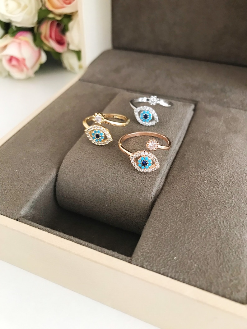 May include: Three adjustable rings with an evil eye design. The rings are made of gold, rose gold, and silver. Each ring has a blue evil eye with a white pupil and is surrounded by clear rhinestones.
