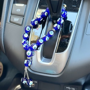 Op de afbeelding: Een blauwe auto-hanger met kralen met meerdere glazen kralen met een oogontwerp. De hanger heeft een zilverkleurige ketting en hangt in een auto. De kralen zijn diepblauw met witte en lichtblauwe oogdetails.