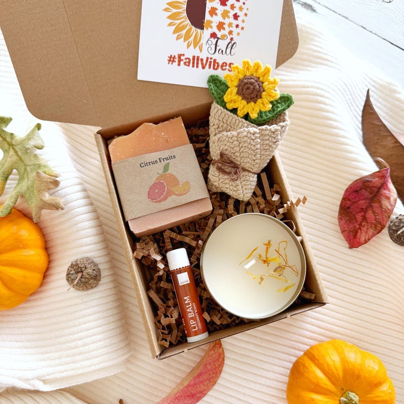 May include: A gift box containing a bar of Citrus Fruits soap, a crochet sunflower in a beige wrap, a round candle with dried petals, and a tube of lip balm. A card with the text Fall #FallVibes is tucked into the open lid of the cardboard box.