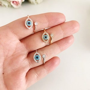 May include: Three adjustable rings with an evil eye design. Each ring features a blue evil eye with a white and black pupil surrounded by clear rhinestones. The rings are made of silver, gold, and rose gold.