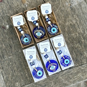 May include: Six small white boxes with blue and gold evil eye charms hanging from white string with blue and white beads. The boxes have white labels with the text "The Evil Eye" and a description of the charm.