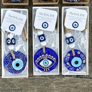 Puede incluir: Tres cajas blancas con amuletos de ojo turco azules y blancos en su interior. Cada caja tiene una tarjeta blanca con el texto "The Evil Eye" y una descripción del amuleto. Los amuletos están en una cuerda con cuentas blancas y cuentas de ojo turco azules.
