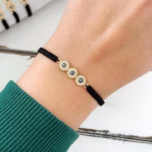May include: A black string bracelet with three gold evil eye charms. Each charm is decorated with white stones and a blue eye.