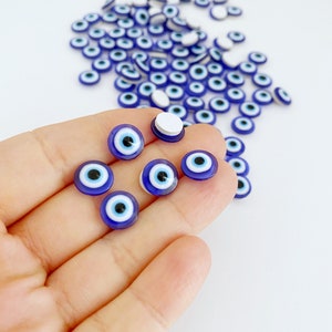 May include: A close-up of a hand holding a group of small blue and white evil eye charms. The charms are round and have a white center with a blue circle around it. There are many more charms scattered on a white surface.