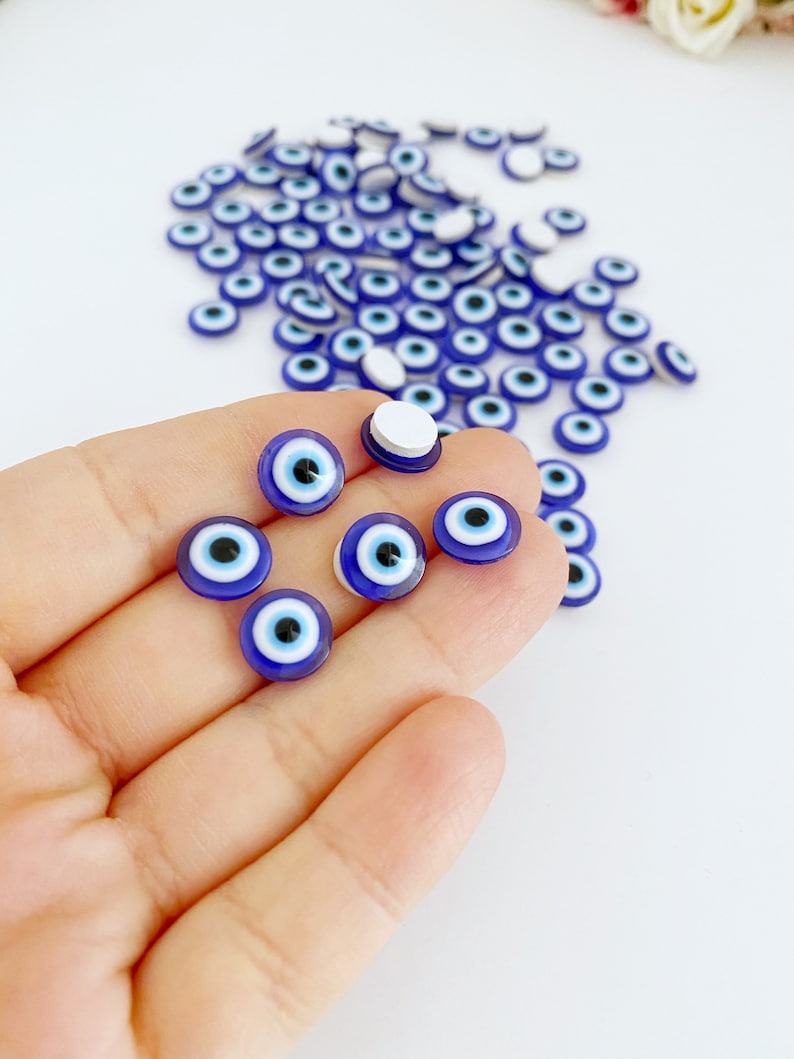 May include: A close-up of a hand holding a group of small blue and white evil eye charms. The charms are round and have a white center with a blue circle around it. There are many more charms scattered on a white surface.