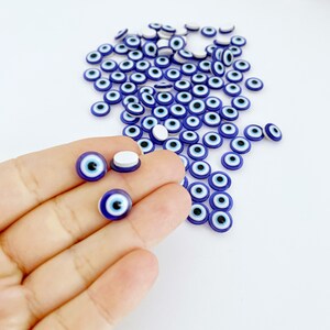 May include: A large quantity of small blue and white evil eye charms. The charms are round and have a white center with a black pupil. The charms are scattered on a white surface.