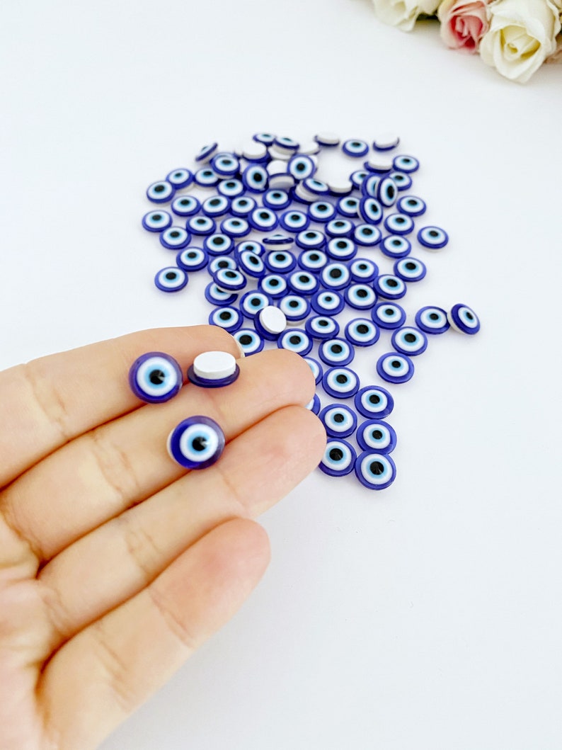 May include: A large quantity of small blue and white evil eye charms. The charms are round and have a white center with a black pupil. The charms are scattered on a white surface.