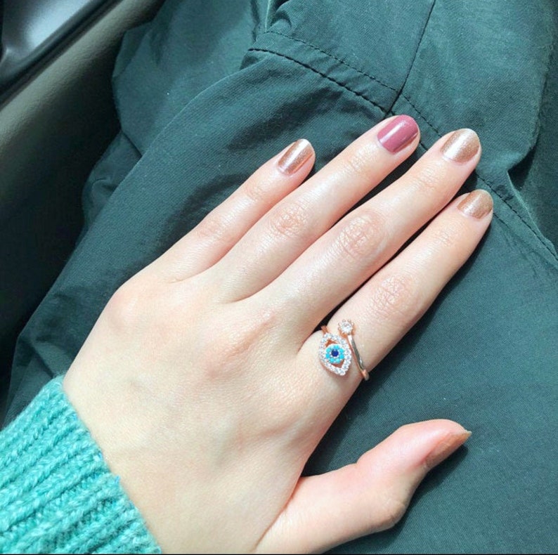 May include: A hand wearing a gold ring with a blue and white evil eye design. The ring is on the middle finger of the hand. The hand is also wearing nail polish in various shades of pink and nude.