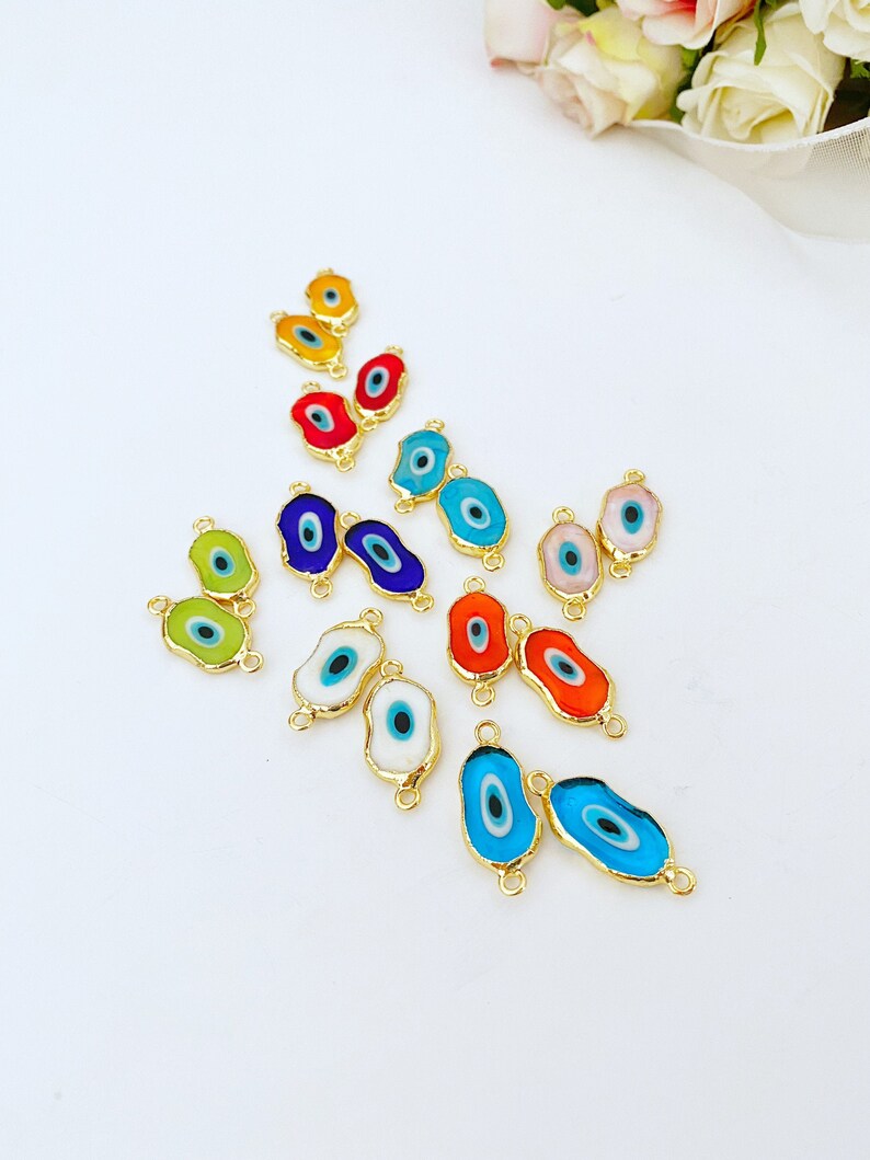 May include: Assortment of colorful evil eye charms with gold accents. The charms are shaped like an eye and have a blue pupil. They are arranged in a pattern on a white background.