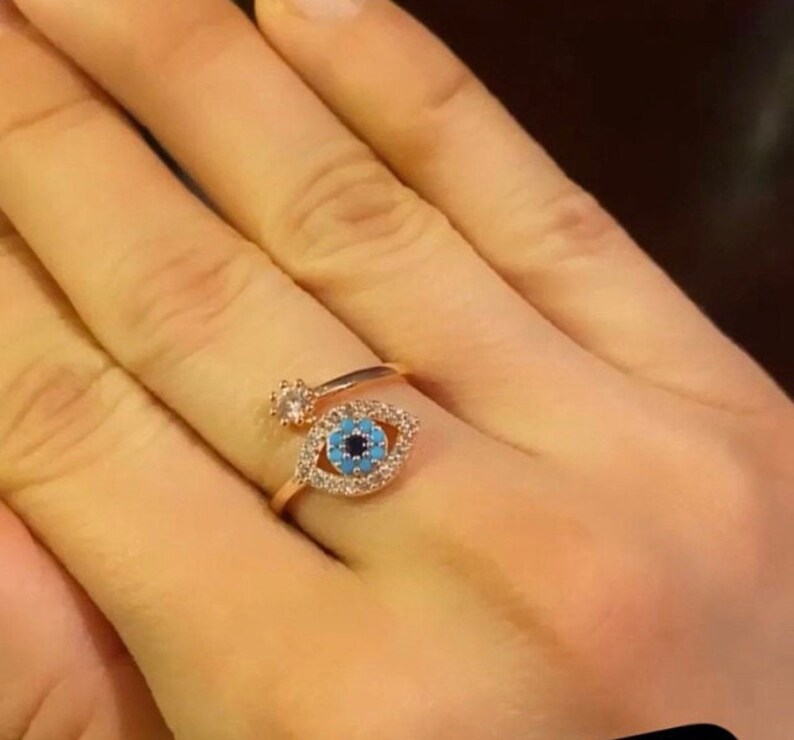 May include: A gold ring with a blue evil eye charm surrounded by clear rhinestones. The ring is on a finger.