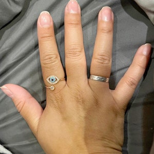 May include: A hand wearing two rings. The ring on the index finger is a gold ring with a blue evil eye design. The ring on the middle finger is a silver band.