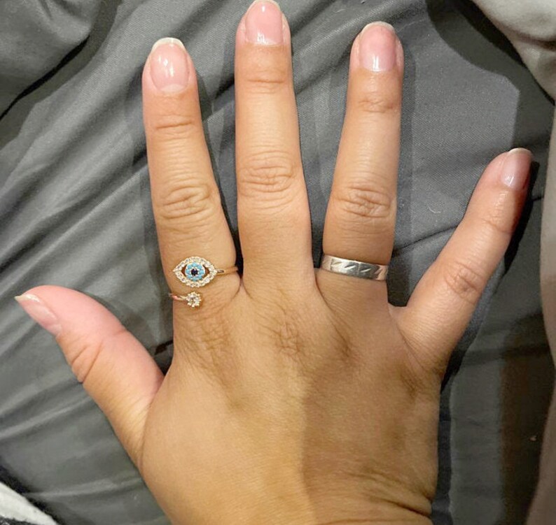 May include: A hand wearing two rings. The ring on the index finger is a gold ring with a blue evil eye design. The ring on the middle finger is a silver band.