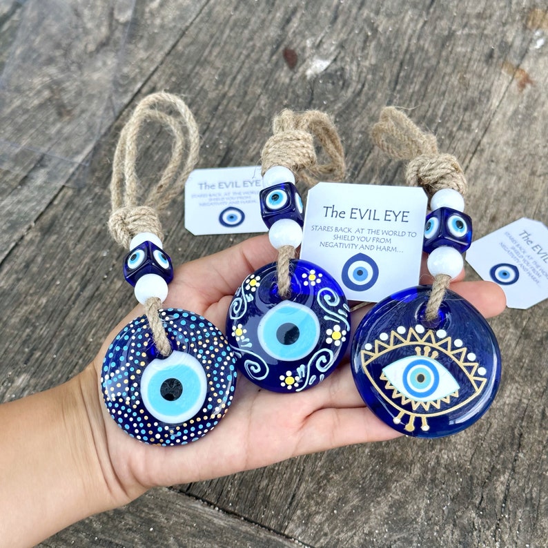 May include: Three blue glass evil eye charms with white and gold accents. The charms are hanging from twine with white beads and blue evil eye beads. The charms are labelled "The Evil Eye" and have text that reads "Stares back at the world to shield you from negativity and harm."