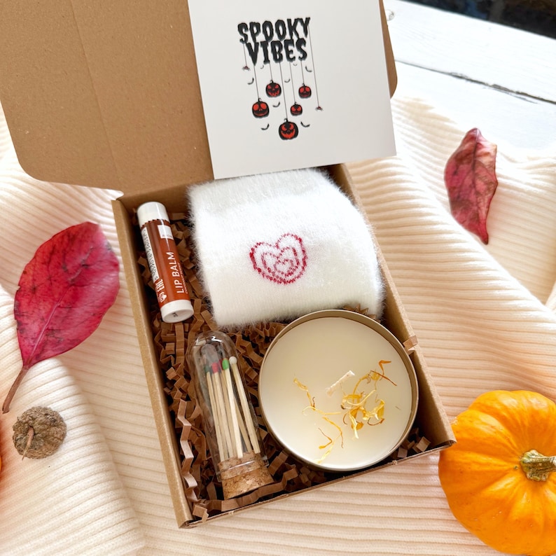 May include: A cardboard gift box containing a pair of white fuzzy socks with a red heart, a tube of lip balm, a glass vial of matches with colored tips, and a round candle with dried petals. A card inside reads SPOOKY VIBES with pumpkin art.