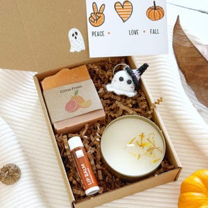May include: A gift box containing a bar of Citrus Fruits soap, a white lip balm, a round candle with dried petals, and a small crocheted ghost keychain wearing a black hat. A card inside reads Peace, Love, Fall with icons of a hand, heart, and pumpkin.
