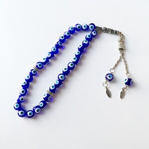 Puede incluir: Un collar de cuentas de oraci&oacute;n de ojo turco azul y blanco con detalles plateados y dos amuletos de ojo turco con hojas plateadas.