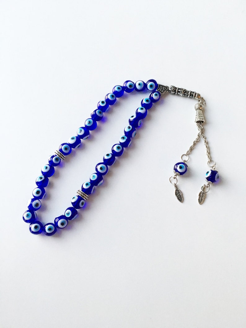 Puede incluir: Un collar de cuentas de oraci&oacute;n de ojo turco azul y blanco con detalles plateados y dos amuletos de ojo turco con hojas plateadas.