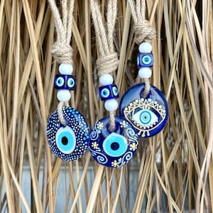 May include: Three blue and white evil eye charms hanging from twine cords. The charms are decorated with gold paint and have different patterns.