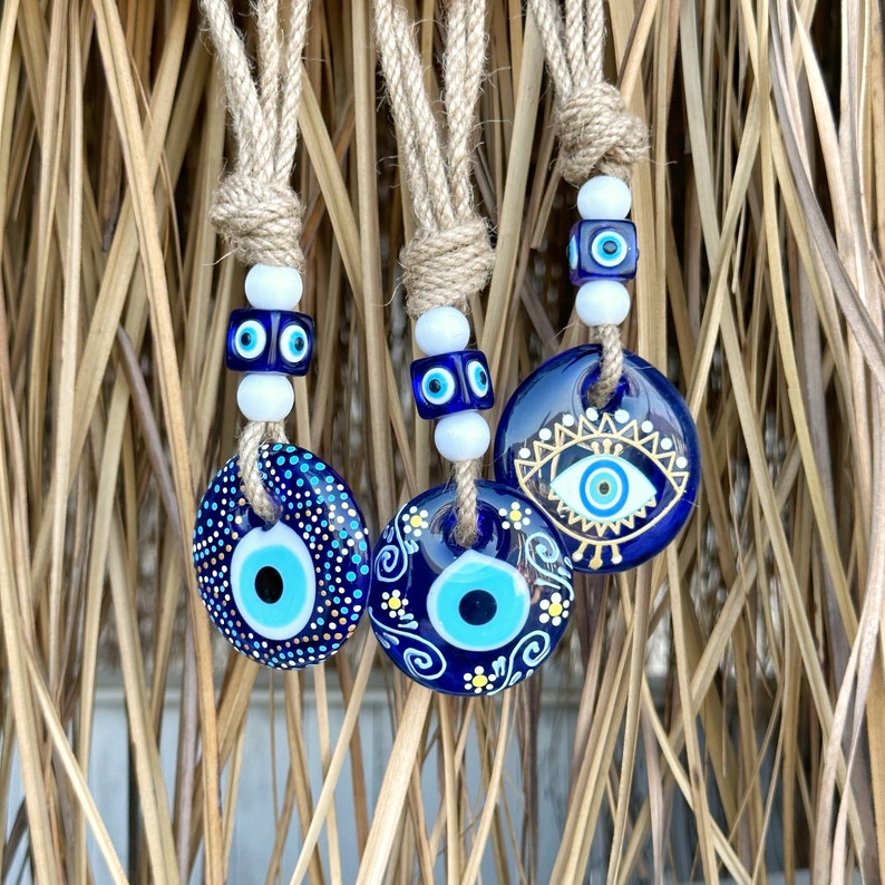 May include: Three blue and white evil eye charms hanging from twine cords. The charms are decorated with gold paint and have different patterns.