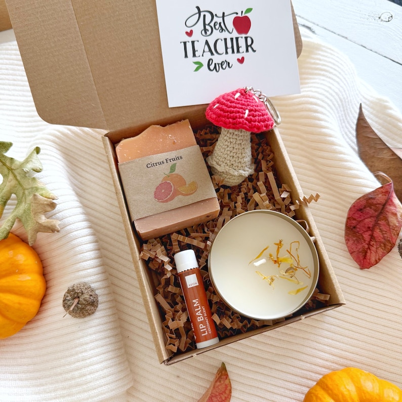 May include: A gift box containing a bar of Citrus Fruits soap with a brown paper label, a red and beige crocheted mushroom keychain, a small round candle with dried petals, and a lip balm tube. A card inside reads Best Teacher Ever.