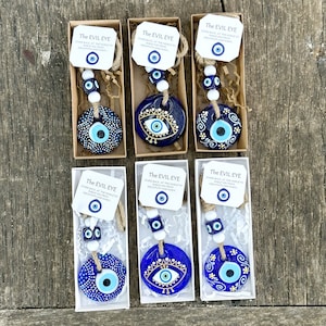 May include: Six small, white boxes with blue and gold evil eye charms hanging from twine. Each box has a small tag with the text "The Evil Eye" and a smaller evil eye charm. The text on the tag reads "Stares back at the world to shield you from negativity and harm."