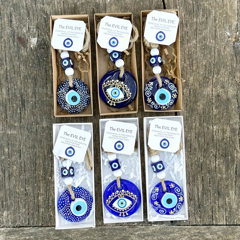 May include: Six small, white boxes with blue and gold evil eye charms hanging from twine. Each box has a small tag with the text "The Evil Eye" and a smaller evil eye charm. The text on the tag reads "Stares back at the world to shield you from negativity and harm."
