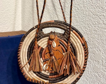 May include: Round woven handbag with a braided leather strap and handle. The bag features a geometric design with natural, brown, and black tones. Tassels and decorative leather accents with shells add detail to the bag.