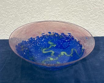 May include: A decorative glass bowl with a wide, shallow design. The rim is a soft pink, transitioning to a vibrant blue interior with green and yellow accents. The bowl is likely used for display or serving.
