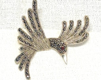 May include: A silver-toned bird brooch with intricate detailing. The bird's body and wings are adorned with small, dark stones, and a single red stone serves as an eye. The beak is pointed, and the brooch is set against a white background.