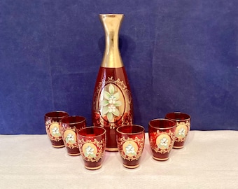 May include: A vintage red glass decanter set with six matching glasses. The decanter has a gold-colored neck and a painted floral design. The glasses and decanter feature a floral design with gold accents. The set is likely for serving a beverage.