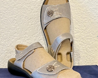 May include: Silver sandals with adjustable straps and a decorative button detail. The sandals have a black sole and a neutral-colored footbed. The sandals are displayed on a blue surface.