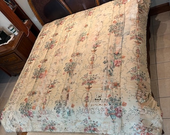 May include: A quilted bedspread featuring a cream-colored background with a repeating pattern of floral bouquets in urns, accented by green vines and pink flowers. The fabric has vertical stitched lines and covers a bed with a dark brown headboard.