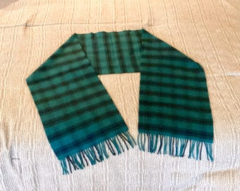 May include: A green and black plaid scarf with fringe at the ends. The scarf is made of a soft material and is laid out on a white textured surface. The scarf has a classic plaid pattern.