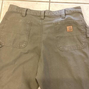 Puede incluir: Vista trasera de pantalones de trabajo verde oliva con m&uacute;ltiples bolsillos y trabillas para el cintur&oacute;n. Un parche de cuero marr&oacute;n con un logotipo es visible en el bolsillo trasero. Los pantalones est&aacute;n hechos de una tela duradera.