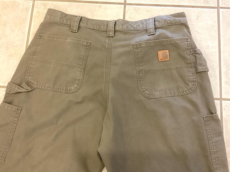 Puede incluir: Vista trasera de pantalones de trabajo verde oliva con m&uacute;ltiples bolsillos y trabillas para el cintur&oacute;n. Un parche de cuero marr&oacute;n con un logotipo es visible en el bolsillo trasero. Los pantalones est&aacute;n hechos de una tela duradera.