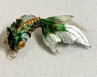 May include: A gold-tone brooch shaped like a fish with green and white enamel scales and fins. The fish features prominent gold-tone eyes with blue centers, a small gold ring hanging from its mouth, and intricate gold-tone detailing along the body.
