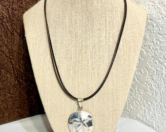 May include: A silver pendant shaped like a sand dollar featuring a textured star pattern in the center, suspended from a thin, dark brown cord necklace. The circular pendant has a polished metallic finish and hangs from a simple silver bail.