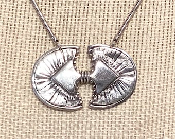 May include: Silver-toned necklace featuring a pendant composed of two semi-circular halves that appear to be joined. Each half has a textured, fan-like design radiating from a central square. The necklace chain is a thin, snake-like style.