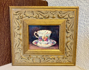 May include: A framed painting of a teacup and saucer with a fruit design. The teacup and saucer are white with colorful fruit illustrations. The frame is ornate, with a gold and cream color scheme.