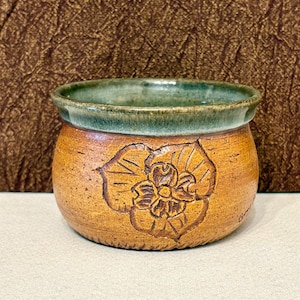 May include: A small, round ceramic bowl with a textured, golden-brown exterior featuring a carved floral design on the front. The interior and the upper rim are glazed in a deep, mottled green, creating a distinct contrast with the warm base.