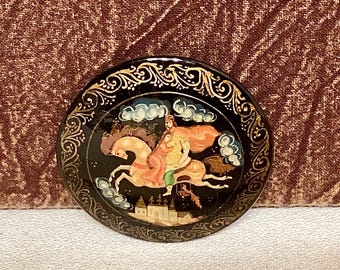 May include: A round black and gold painted brooch with a woman riding a white horse. The woman is wearing a yellow and red dress and a crown. The background is black with gold swirls and clouds.