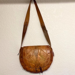 May include: A brown leather satchel bag with a patchwork design. The bag has a long, adjustable strap and a flap closure with a button.